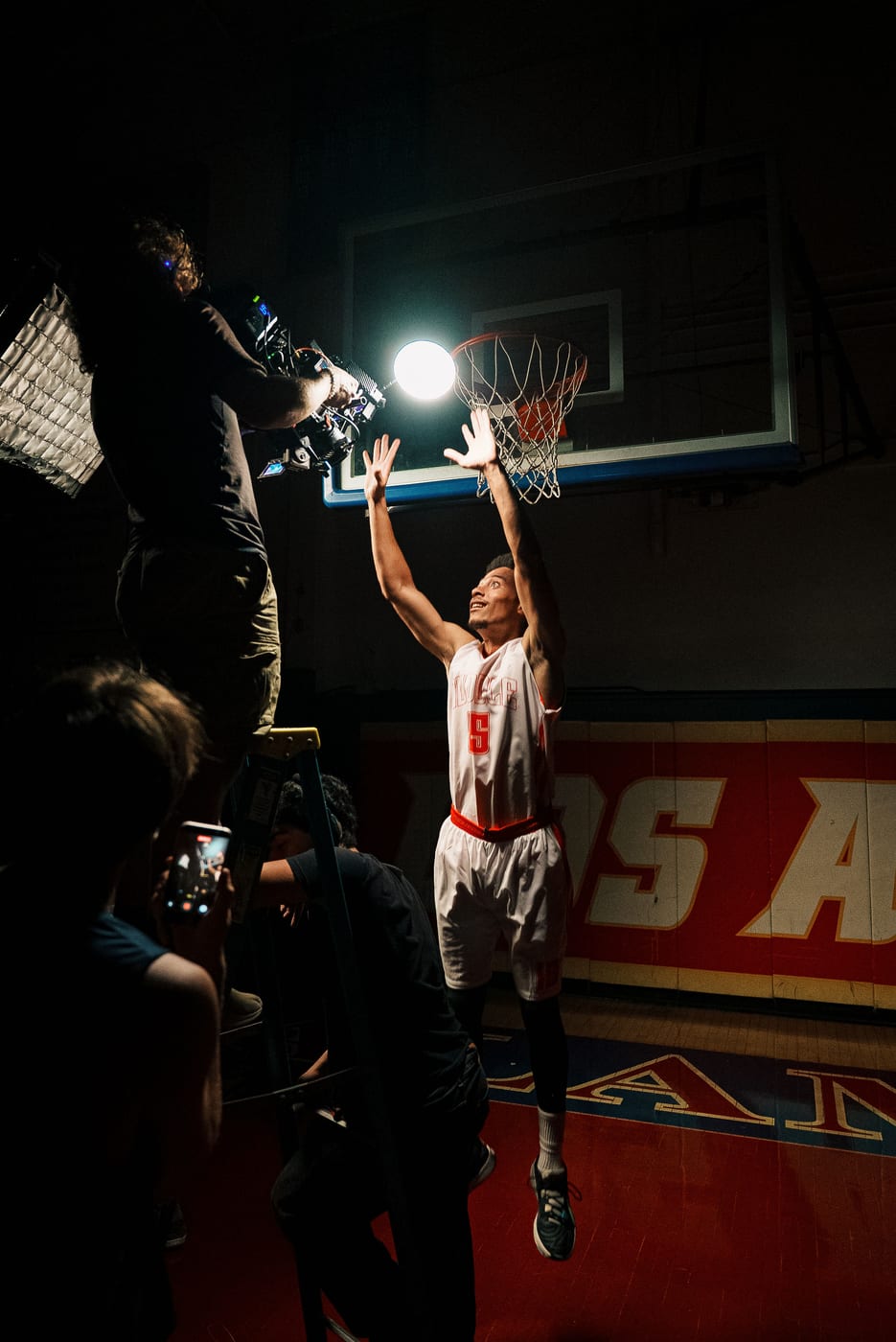 Basketball player reaching toward the hoop while a camera operator films from a ladder